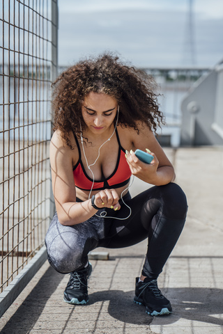 Young athletic woman with smartphone and earbuds crouching outdoors looking at wristwatch stock photo