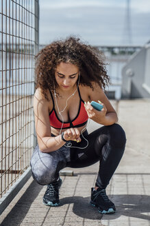 Junge sportliche Frau mit Smartphone und Ohrstöpseln, die im Freien hockt und auf ihre Armbanduhr schaut - VPIF00790