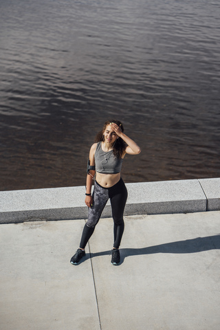 Erschöpfte junge sportliche Frau mit Kopfhörern am Flussufer stehend, lizenzfreies Stockfoto