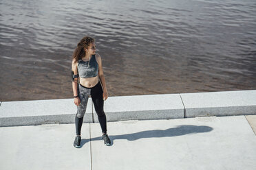 Young athletic woman wearing earbuds standing at the riverside - VPIF00797