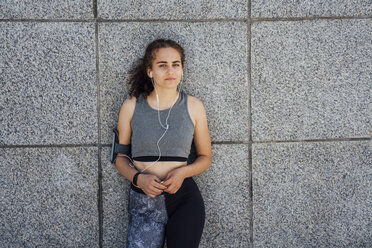 Portrait of young athletic woman wearing earbuds leaning against a wall - VPIF00805