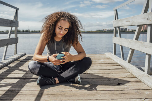 Junge sportliche Frau sitzt am Flussufer und benutzt ihr Smartphone - VPIF00814