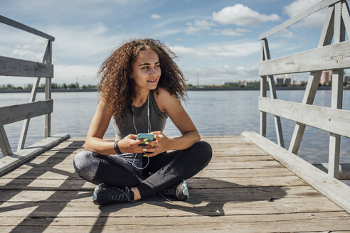 Junge sportliche Frau sitzt am Flussufer und benutzt ihr Smartphone - VPIF00815