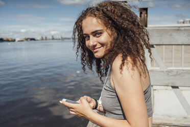 Portrait of smiling young athletic woman with smartphone at the riverside - VPIF00816