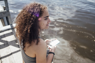 Lächelnde junge sportliche Frau mit Smartphone am Flussufer - VPIF00817