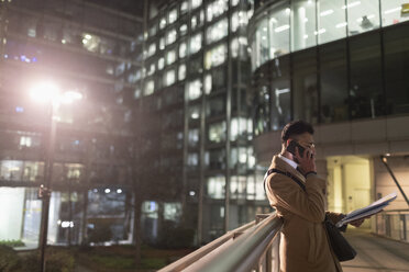 Businessman talking on smart phone, reading paperwork on urban pedestrian bridge at night - CAIF21990