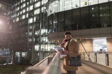 Businessman talking on smart phone and reading paperwork on urban pedestrian bridge at night - CAIF21992