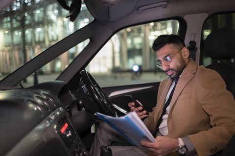 Geschäftsmann mit Smartphone liest Papierkram im Auto bei Nacht, lizenzfreies Stockfoto