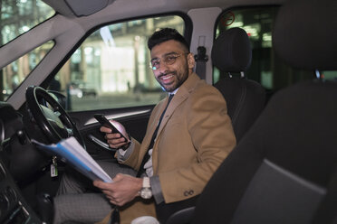 Porträt lächelnder Geschäftsmann mit Smartphone, der nachts im Auto Papierkram liest - CAIF22014