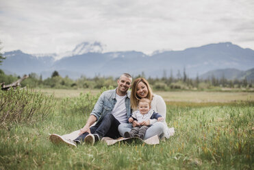 Portrait smiling parents and baby son sitting in rural field with mountains in background - CAIF22023