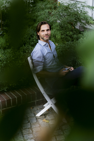 Porträt eines lächelnden Mannes, der auf einem Stuhl im Garten sitzt und ein Mobiltelefon benutzt, lizenzfreies Stockfoto