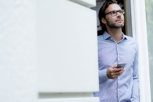 Confident man standing in doorframe holding cell phone - HHLMF00382