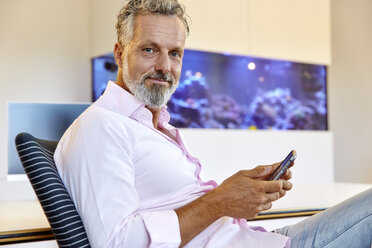 Porträt eines zuversichtlichen Geschäftsmannes, der im Büro sitzt und ein Mobiltelefon benutzt - RHF02136