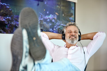 Relaxed mature man listening to music with headphones in front of aquarium - RHF02160