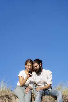 Young couple sitting on a dune in summer, using smartphone - HHLMF00415