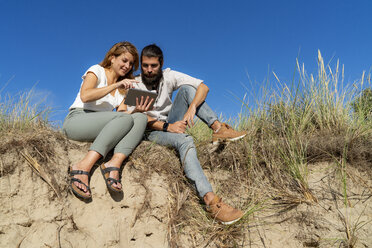 Junges Paar, das im Sommer auf einer Düne sitzt und ein digitales Tablet benutzt - HHLMF00460