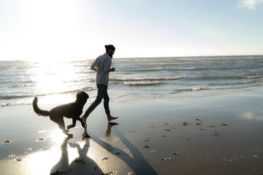 Young man running and playing with his dog on the beach - HHLMF00474