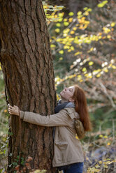 Smiling teenage girl hugging tree trunk in autumnal forest - LBF02047