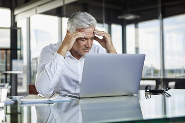 Gestresster Geschäftsmann sitzt mit Laptop am Schreibtisch und hält sich den Kopf - RBF06580