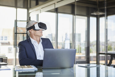 Businessman sitting at desk with laptop, using VR goggles - RBF06599