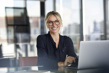 Businesswoman sitting at desk, using laptop, smiling friendly - RBF06640