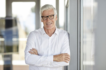 Businessman in office leaning against window, with arms crossed - RBF06645