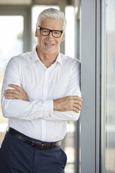 Businessman in office leaning against window, with arms crossed - RBF06648