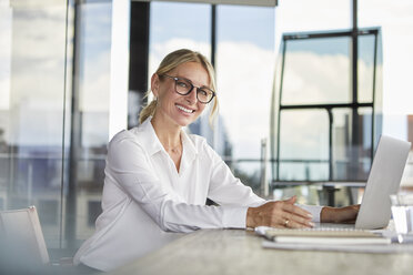 Businesswoman sitting at desk, using laptop, smiling friendly - RBF06705
