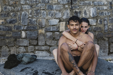 Porträt eines zärtlichen jungen Paares in Badekleidung, das an einer Steinmauer sitzt, lizenzfreies Stockfoto