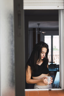 Seriöse schöne junge Frau mit einer Tasse Kaffee am Fenster zu Hause - KKAF01815