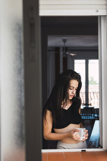 Serious beautiful young woman with cup of coffee at the window at home - KKAF01815