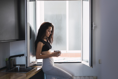 Seriöse schöne junge Frau mit einer Tasse Kaffee am Fenster zu Hause, lizenzfreies Stockfoto