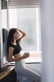 Schöne junge Frau mit einer Tasse Kaffee am Fenster zu Hause - KKAF01817