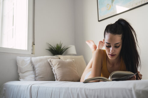 Beautiful young woman lying on bed at home reading a book - KKAF01820