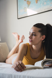 Beautiful young woman lying on bed at home reading a book - KKAF01821