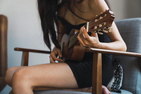 Young woman sitting on armchair at home playing guitar - KKAF01834