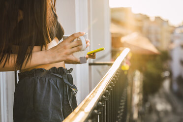 Close-up of young woman on balcony above the city at sunset holding cell phone and cup of tea - KKAF01836