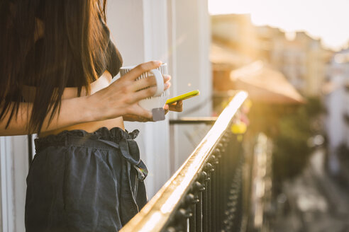 Nahaufnahme einer jungen Frau auf einem Balkon über der Stadt bei Sonnenuntergang mit Handy und Tasse Tee - KKAF01836