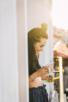 Glückliche junge Frau auf Balkon über der Stadt bei Sonnenuntergang mit Handy und Tasse Tee - KKAF01837