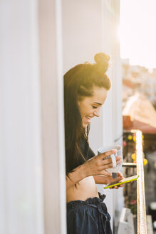 Happy young woman on balcony above the city at sunset holding cell phone and cup of tea - KKAF01837