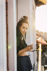 Porträt einer schönen jungen Frau auf einem Balkon über der Stadt bei Sonnenuntergang mit einer Tasse Tee - KKAF01838