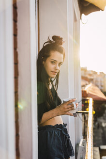 Porträt einer schönen jungen Frau auf einem Balkon über der Stadt bei Sonnenuntergang mit einer Tasse Tee - KKAF01838