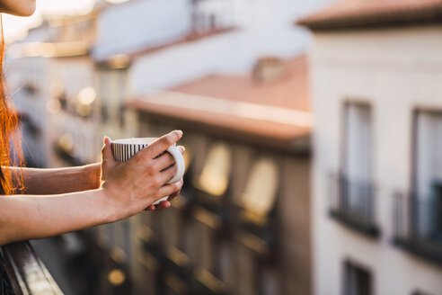 Hände einer Frau auf einem Balkon über der Stadt bei Sonnenuntergang, die eine Tasse Kaffee hält - KKAF01839