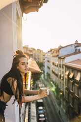 Beautiful young woman on balcony above the city at sunset with cup of coffee - KKAF01840