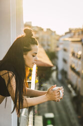 Schöne junge Frau auf Balkon über der Stadt bei Sonnenuntergang mit einer Tasse Kaffee - KKAF01841