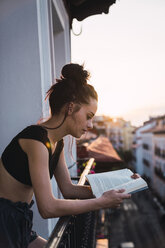 Schöne junge Frau auf Balkon über der Stadt bei Sonnenuntergang liest ein Buch - KKAF01848