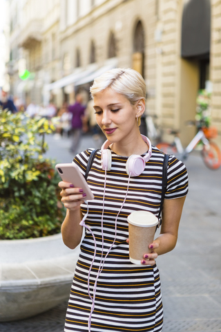 Porträt einer jungen Frau in einem gestreiften Kleid, die mit einem Kaffee auf der Straße steht und auf ihr Mobiltelefon schaut, lizenzfreies Stockfoto