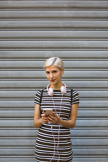 Porträt einer jungen Frau mit Kopfhörern und Smartphone in einem gestreiften Kleid vor einem Rolltor - MGIF00250