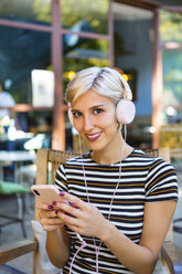 Porträt einer lächelnden jungen Frau mit Smartphone, die mit Kopfhörern in einem Straßencafé Musik hört - MGIF00252