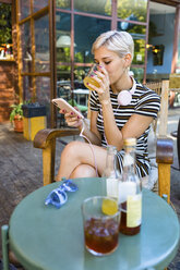 Junge Frau in einem Straßencafé, die ein Erfrischungsgetränk genießt und auf ihr Smartphone schaut - MGIF00255